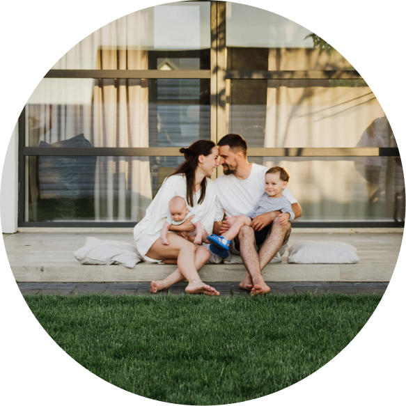 A family of four sitting closely on a porch with green grass in the foreground, smiling and holding their two young children.