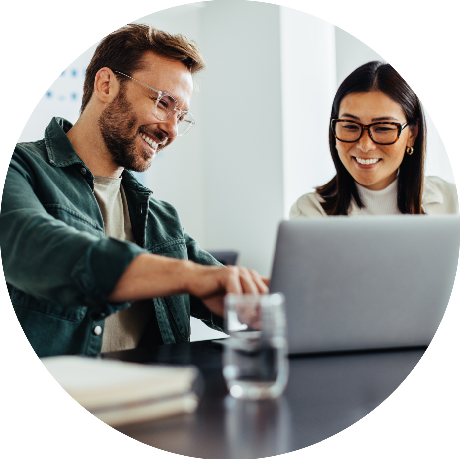Man and woman wearing glasses smiling while working on a laptop together at a desk.