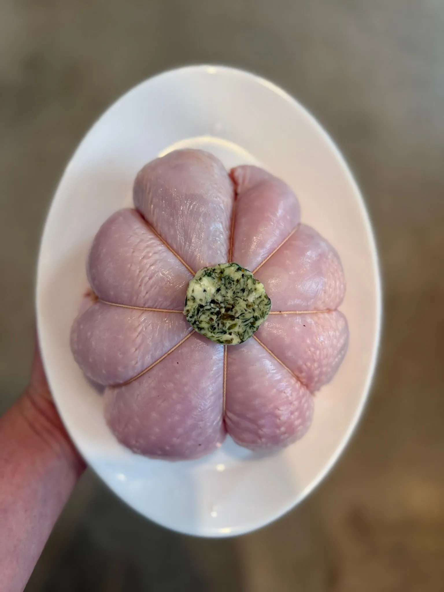 Raw chicken wrapped with string on a white oval plate with herb butter on top.