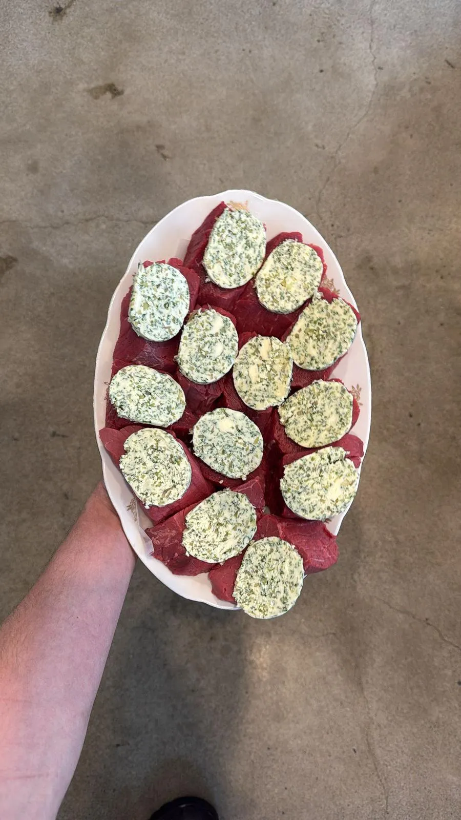 Hand holding a plate of raw beef slices topped with herb butter rounds.