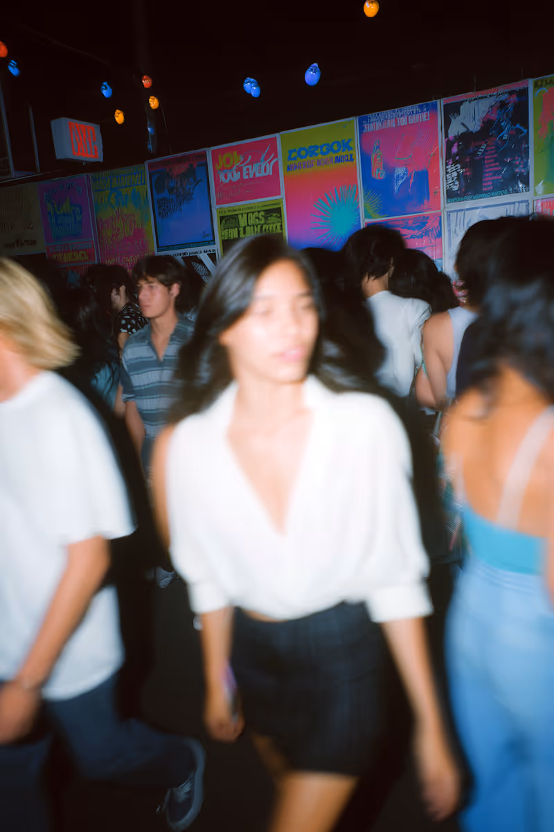 Blurred image of people walking in a dimly lit space with colorful posters on the wall and string lights above.