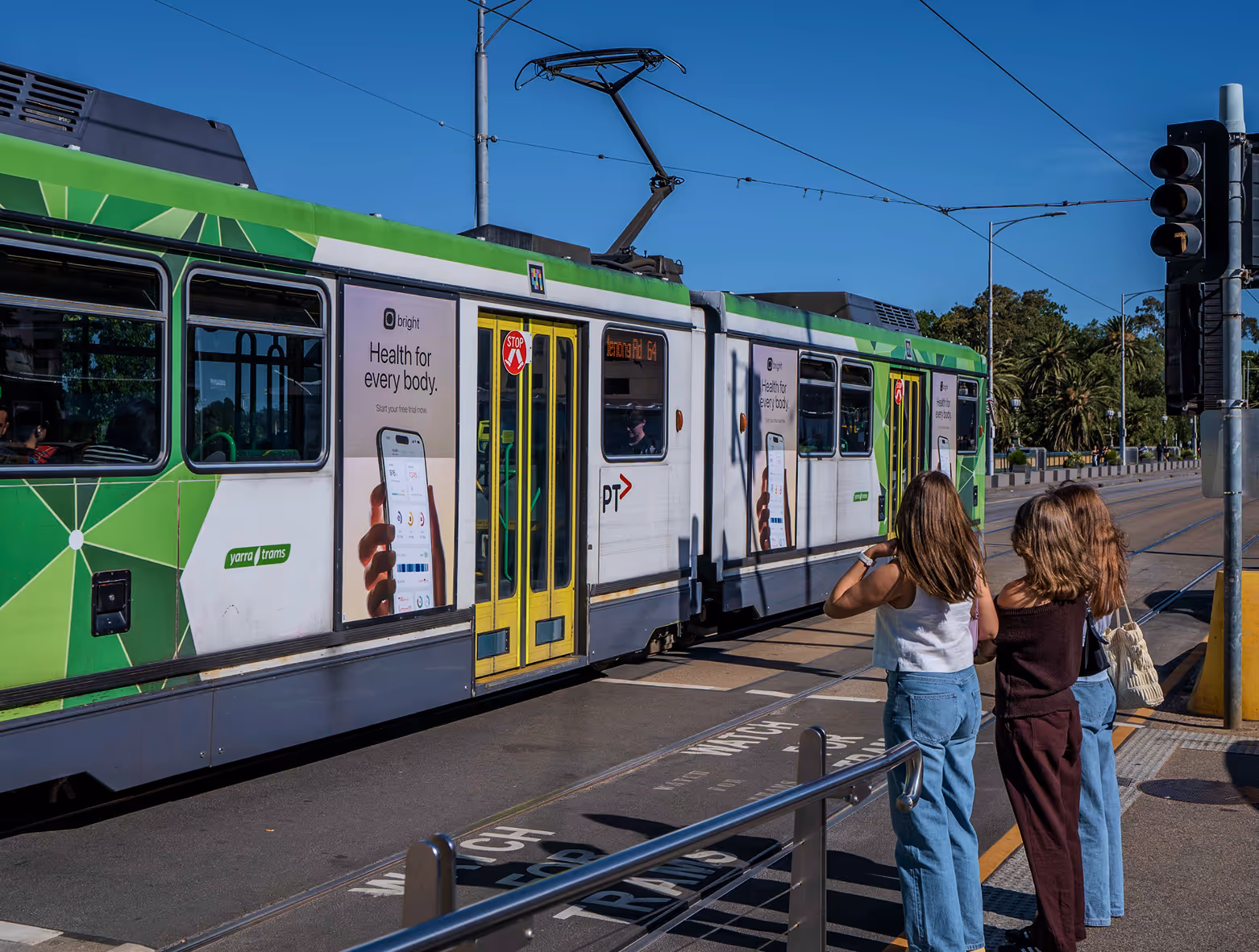 Two girls stand at a tram stop watching a green and white Yarra Trams vehicle with a health app advertisement on its side.