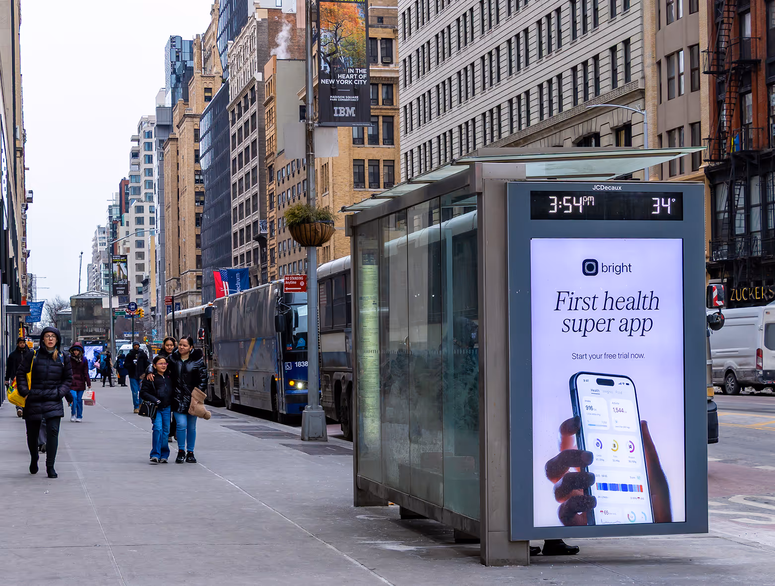 City street scene with a modern bus shelter displaying a digital ad for the Bright health super app, showing the time as 3:54 PM and temperature as 34°F.