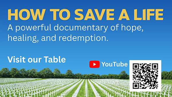 Outdoor scene with two signs near a road; one sign lists community services at Our Lady of the Rosary Catholic Church, the other promotes a YouTube video titled 'How to Save a Life' with graves marked by white crosses on the ground beneath.