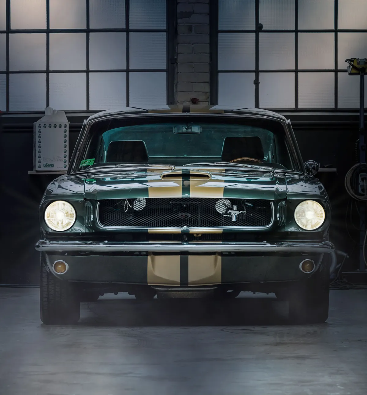 Front view of a 1965 Ford Mustang in dark green with gold racing stripes and headlights on in a garage.