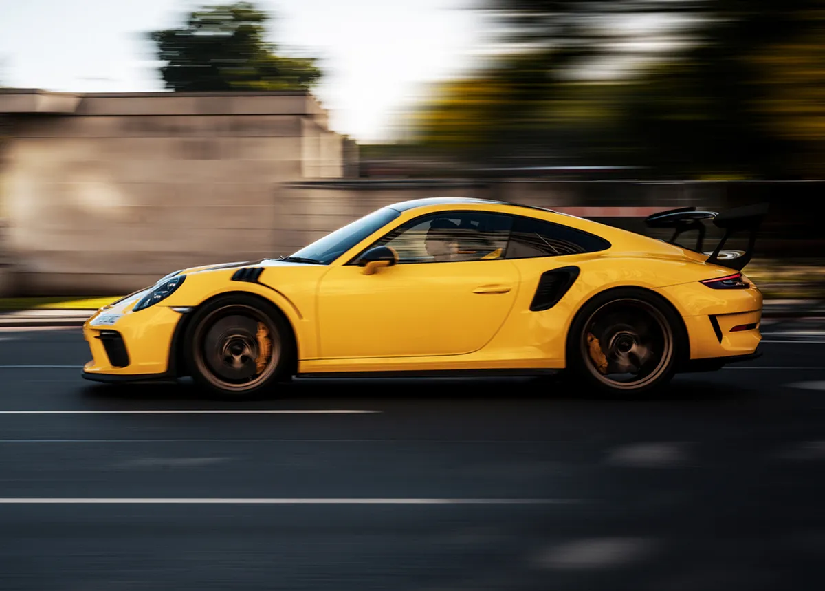 Yellow Porsche 9112 GT2 with black spoiler speeding on road with blurred background of trees and wall.