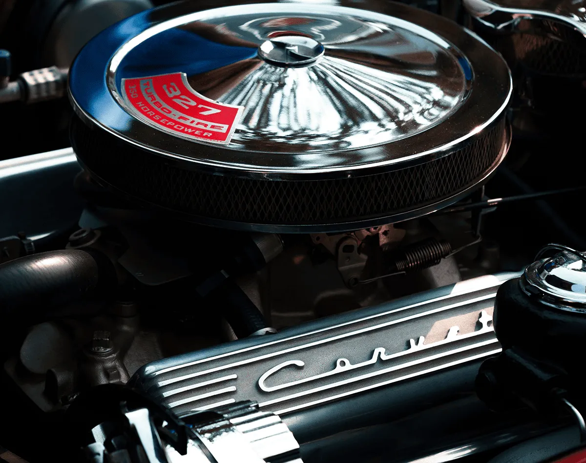 Close-up of a Corvette engine featuring a circular chrome air cleaner with a red decal reading '327 Turbo-Fire 350 Horsepower' and a metal valve cover with the scripted 'Corvette' logo.