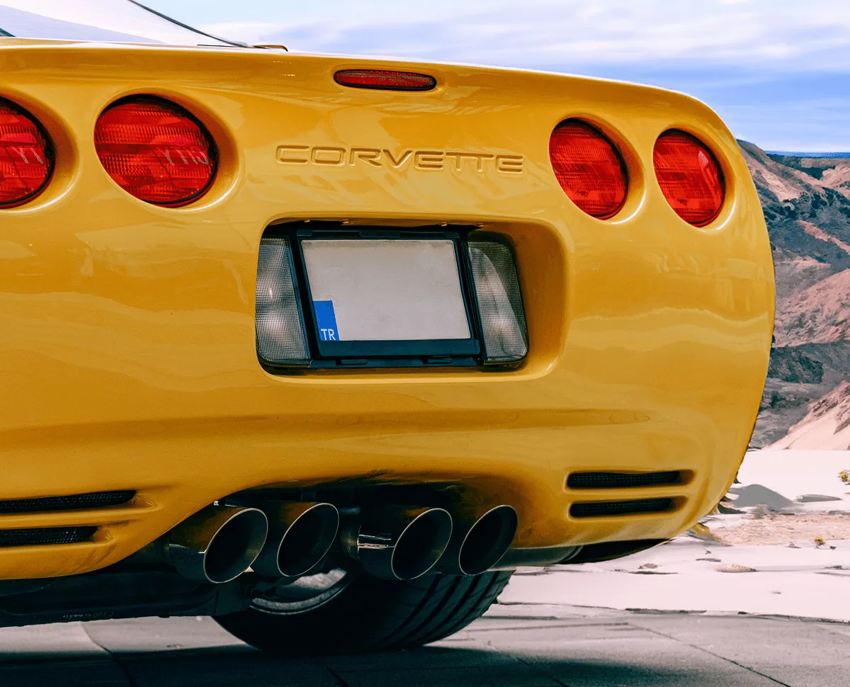 Rear view of a yellow Chevrolet Corvette showing four red circular tail lights, dual exhaust pipes, and a license plate with a blue 'TR' tag.