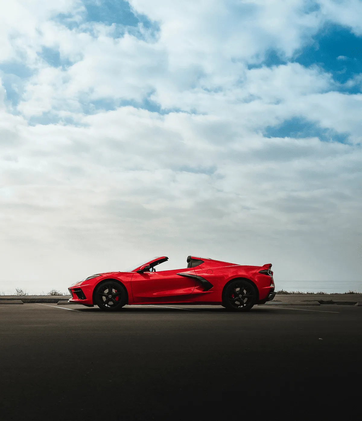 Side view of a red convertible sports car parked in an empty lot under a partly cloudy blue sky.