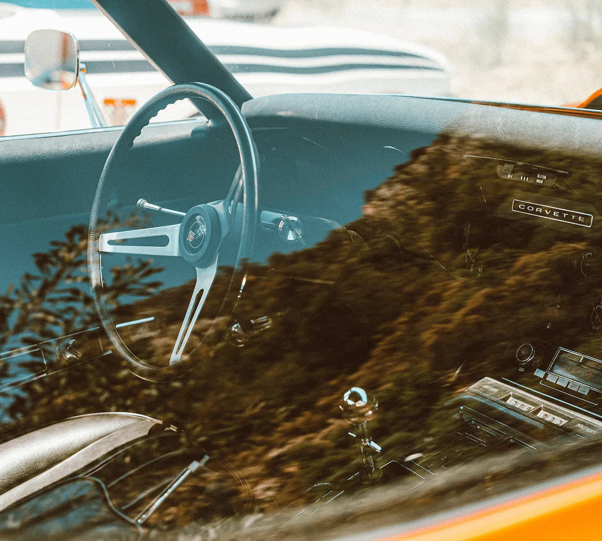 View through the window of a classic Corvette car interior showing the steering wheel, dashboard with the Corvette nameplate, gear shift, and leather seats, with reflections of trees on the glass.