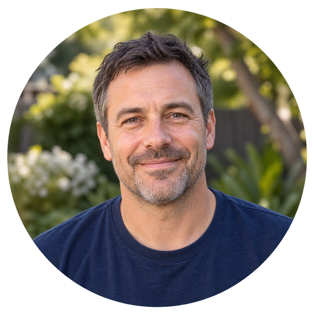Close-up of a middle-aged man with short salt-and-pepper hair and a trimmed beard, wearing a navy blue t-shirt, smiling gently outdoors with blurred green foliage and flowers in the background.
