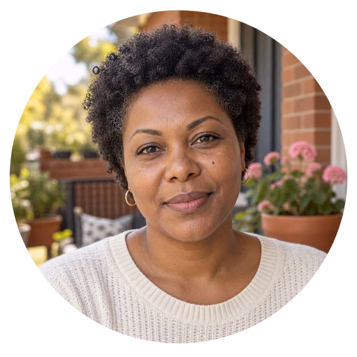 Portrait of a woman with short curly hair wearing a white knit sweater and small hoop earrings, sitting outdoors with flowering plants and a brick wall in the background.