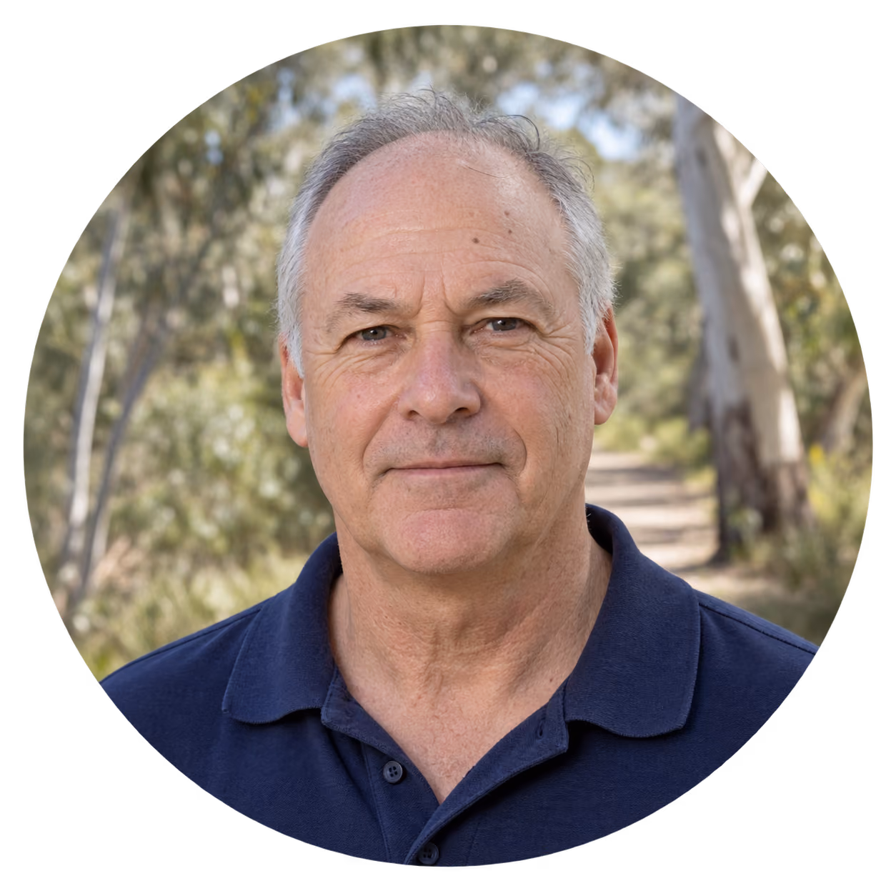 Portrait of a middle-aged man with short gray hair wearing a navy blue polo shirt, standing outdoors with trees and a pathway in soft focus behind him.
