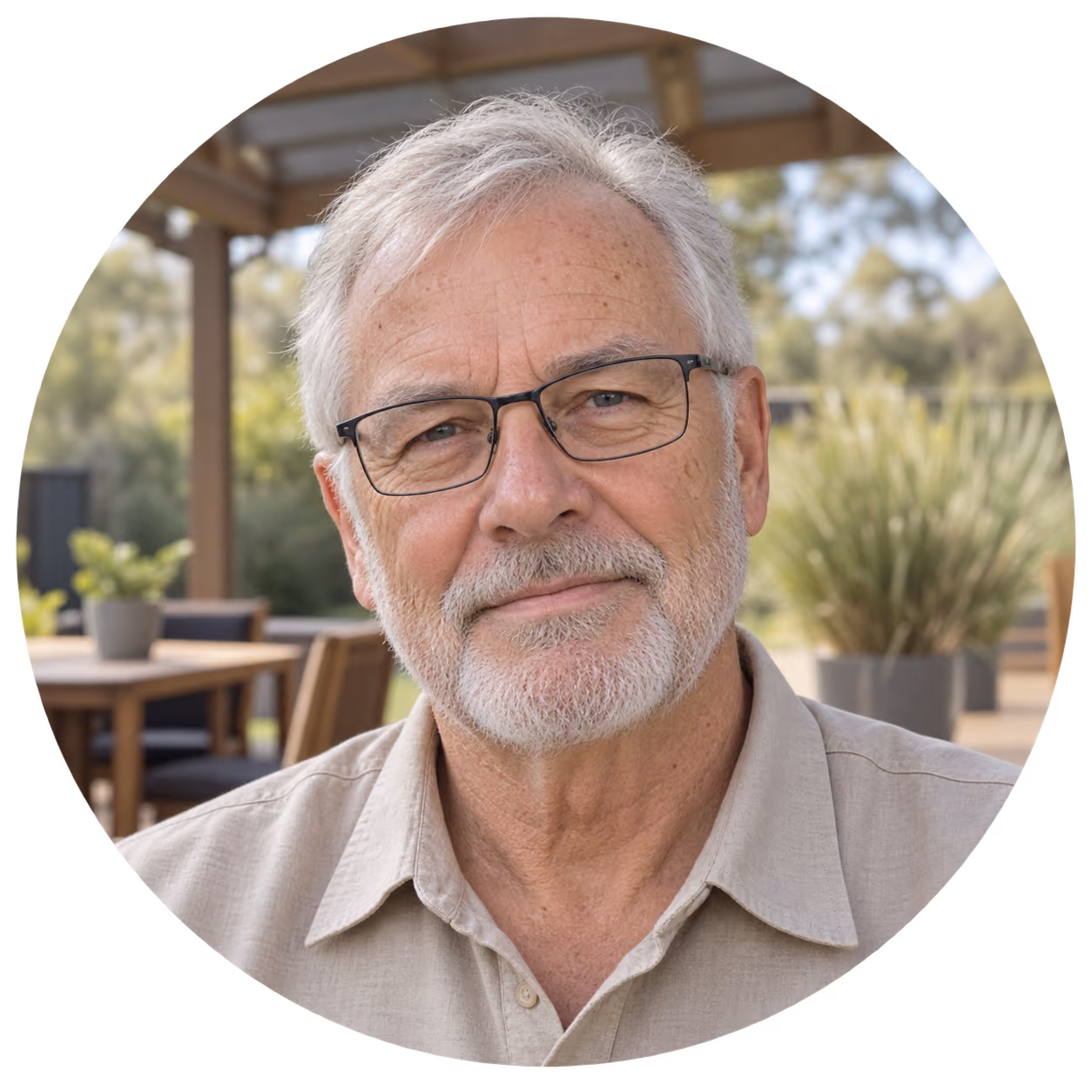 Portrait of an elderly man with gray hair, a beard, and glasses, wearing a beige shirt, sitting outdoors with blurred greenery and wooden furniture in the background.