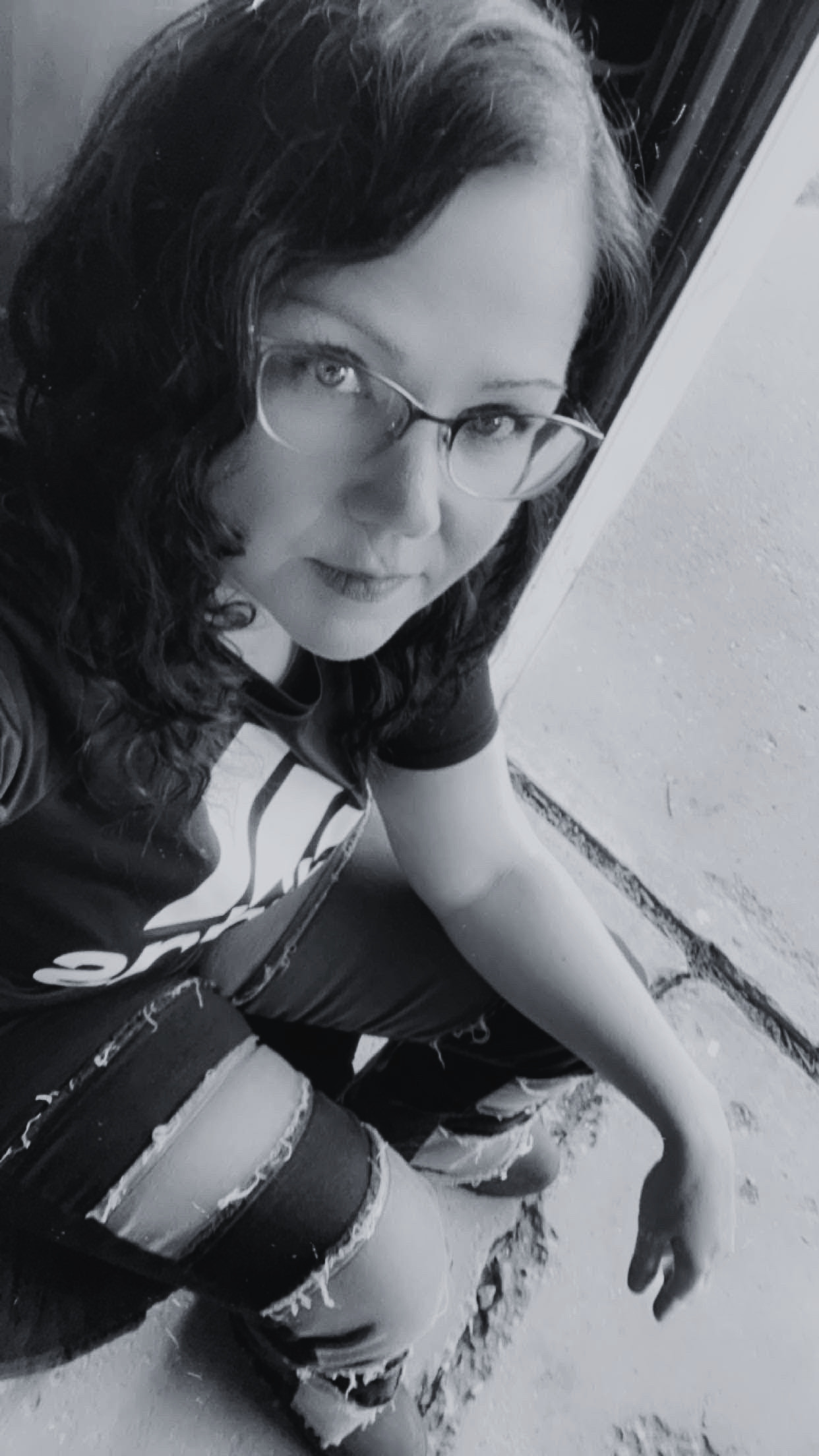 Black and white photo of a woman with glasses and curly hair sitting near a doorway, wearing ripped jeans.