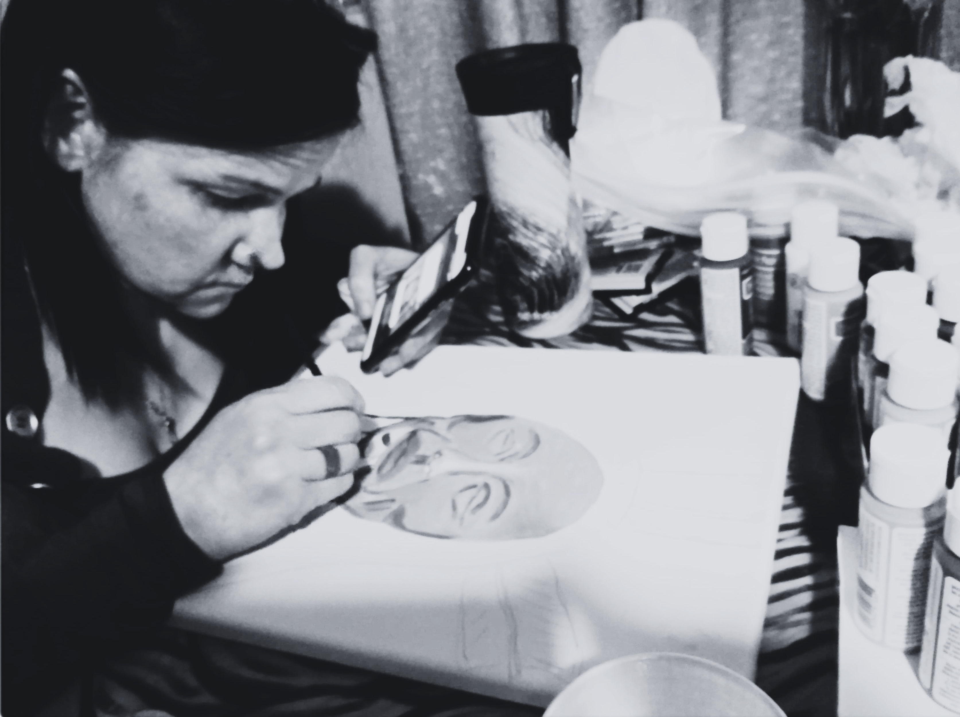 Woman focused on painting a human face on a canvas while holding a phone in her left hand.
