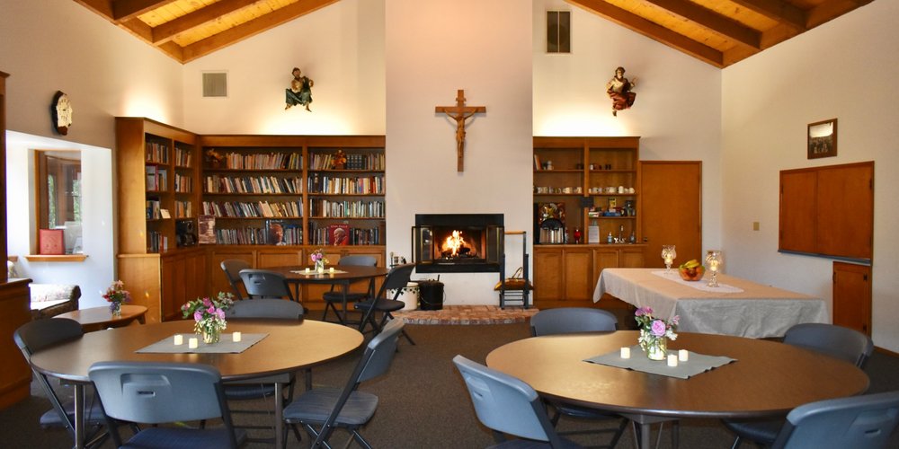 Cozy room with round tables, chairs, floral centerpieces, a lit fireplace with a crucifix above, bookshelves, and wooden ceiling beams.