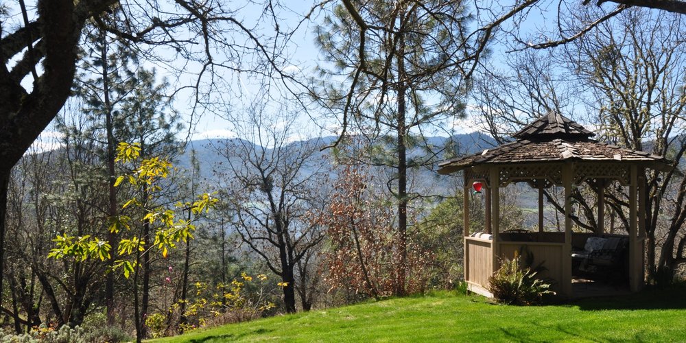 Wooden gazebo on green grass surrounded by trees with mountains visible in the background under a clear sky.