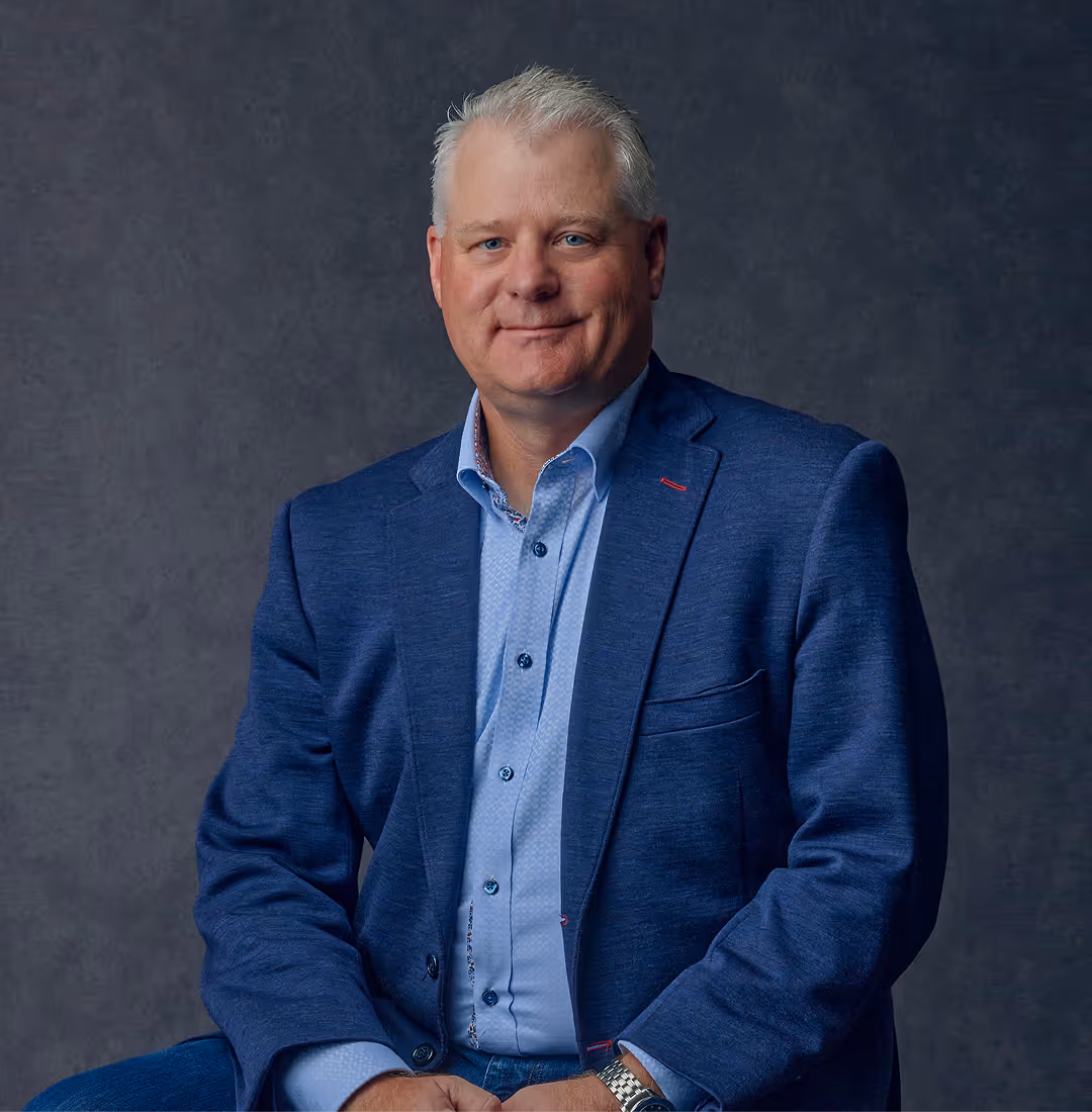 Middle-aged man with short gray hair wearing a blue blazer and light blue shirt sitting against a dark gray background.