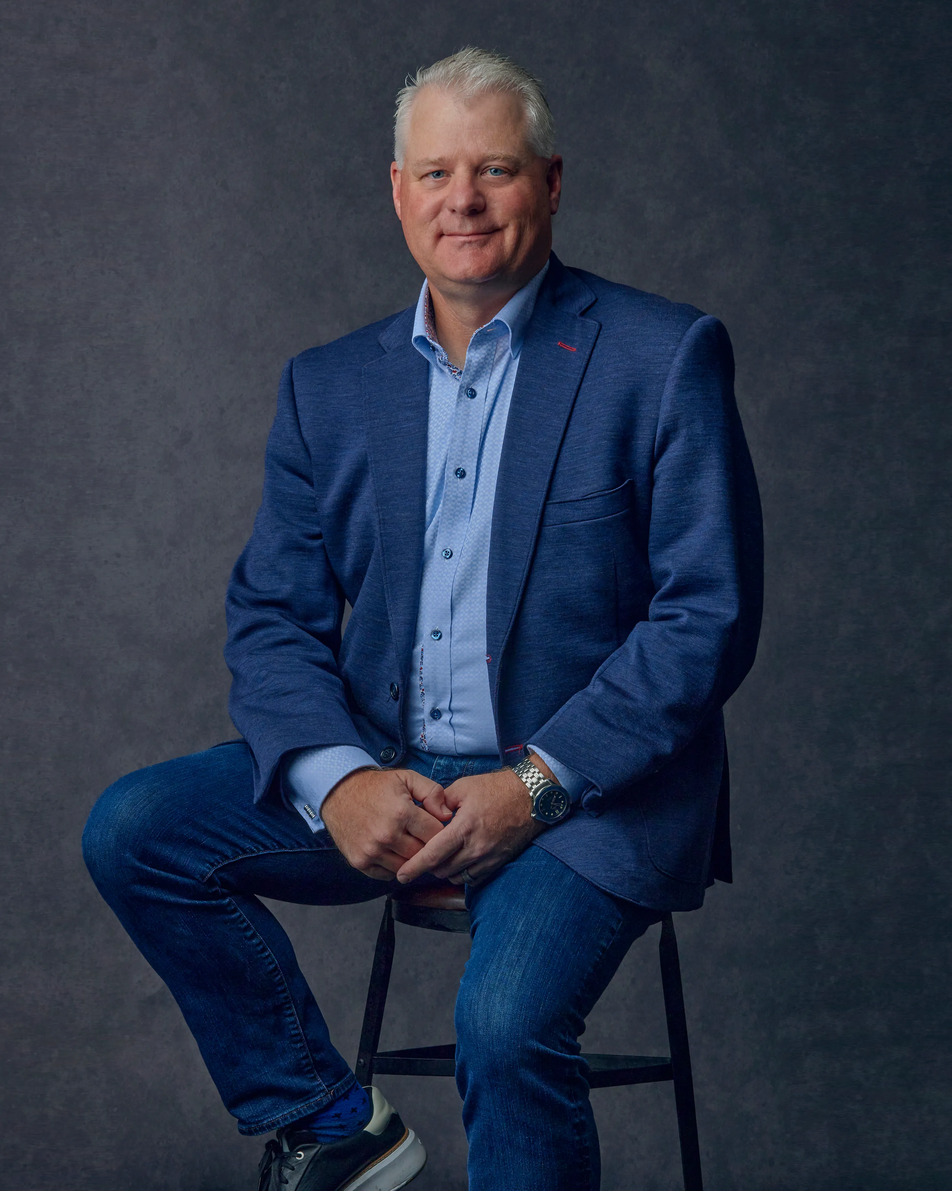 Man with short gray hair wearing a blue blazer, light blue shirt, jeans, and sneakers sitting on a stool against a gray backdrop.