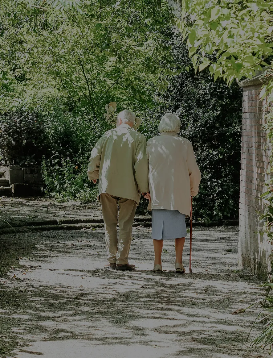 Elderly couple walking hand in hand on a shaded garden path surrounded by greenery.