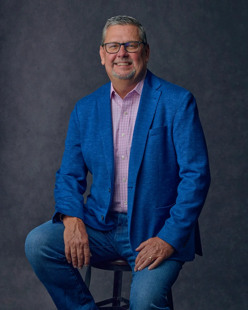Smiling middle-aged man wearing glasses, a blue blazer, pink checkered shirt, and jeans, sitting on a stool against a dark background.