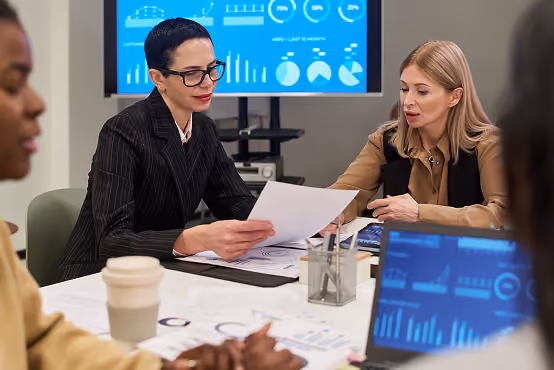 Three professionals in a meeting reviewing documents and data charts on a screen and laptop.