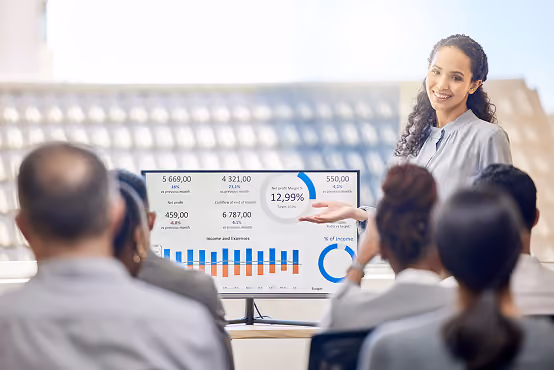 Smiling woman presenting financial charts and graphs to an audience in a meeting room.