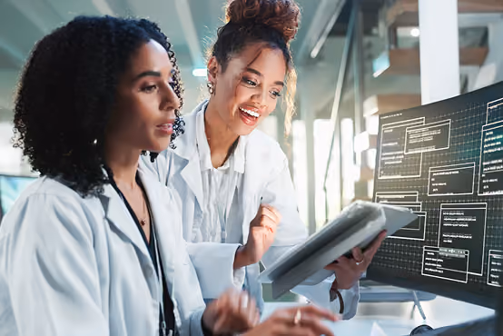 Two women scientists in lab coats discuss data shown on a digital screen and tablet in a modern lab.