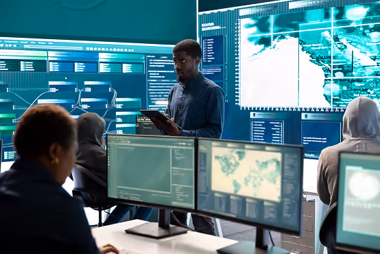 Man with tablet stands in front of large digital maps and data screens with people working at computers.