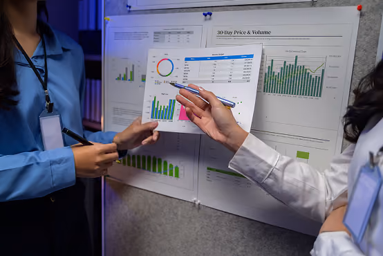 Two people discuss graphs and charts on a bulletin board, one pointing at a chart with a pen.