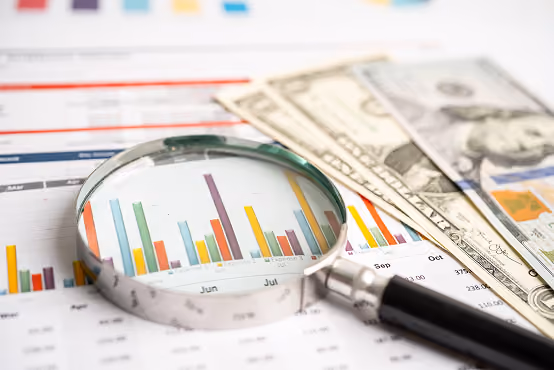 Magnifying glass over colorful bar charts with US dollar bills on financial papers.
