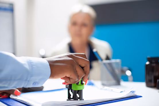 Hand stamping a document with a woman blurred in the background during an office meeting.