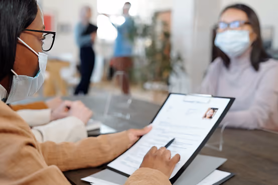 Two masked people sit across a desk with a clear partition, one reviewing a resume or document.