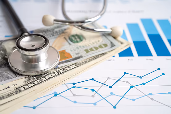 Stethoscope on US dollar bills overlaid on financial charts with blue and gray line graphs.