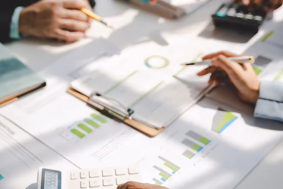 Hands pointing at charts and graphs on paper, with a calculator and pen on a desk.