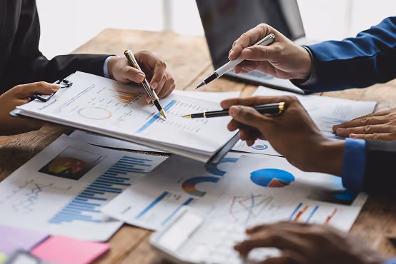 Multiple hands holding pens pointing at business charts and graphs on paper at a wooden table.