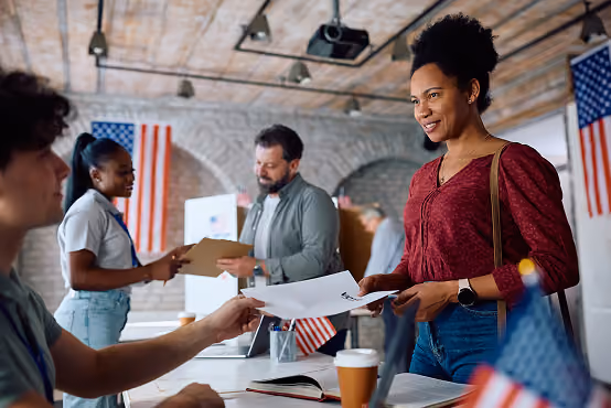 Woman handing documents to seated man in office with American flags and coworkers in background.