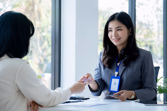 Smiling woman in business attire hands a card to another woman across a desk by a window.