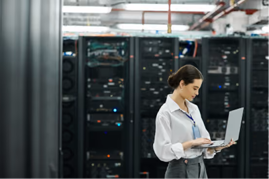 Woman in data center working on a laptop near server racks and technology equipment.