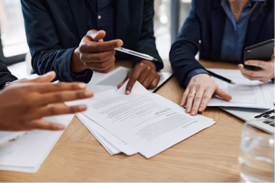 People review and discuss documents at a table during a meeting, one holding a pen and another a phone.