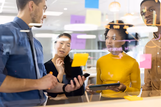 Four colleagues brainstorming in an office behind a glass wall with colorful sticky notes.