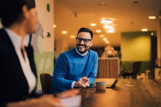 Man in blue sweater smiling and talking with a woman at a wooden desk with a laptop and mug.
