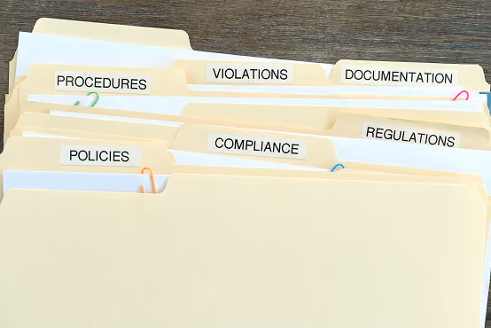 Manila folders labeled Procedures, Violations, Documentation, Policies, Compliance, and Regulations on wooden surface.