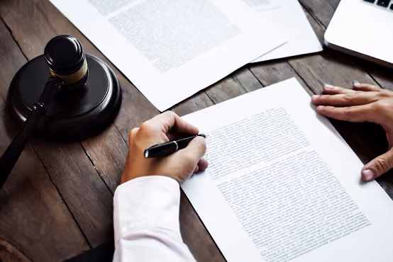 Person signing a legal document on wooden table with a judge's gavel nearby.