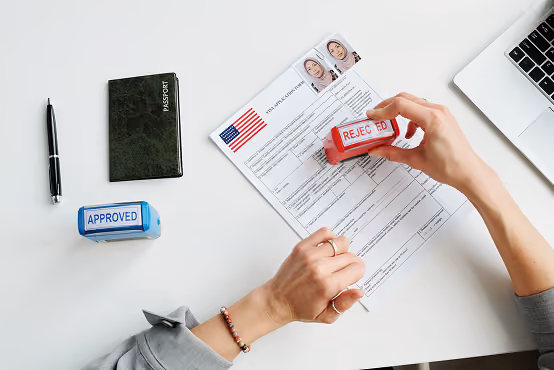 Hands stamping a US visa application with a red REJECTED stamp on a white table with passport and laptop.