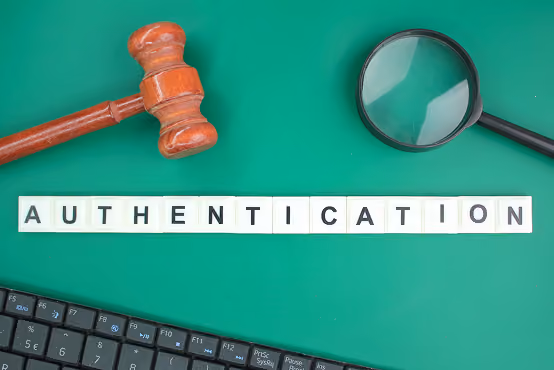 Wooden gavel, magnifying glass, keyboard keys and letter tiles spelling authentication on green surface.