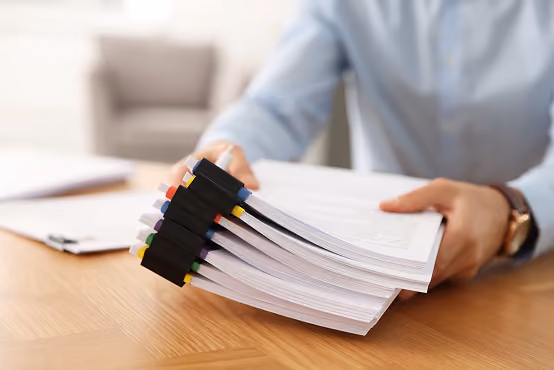 Person holding thick documents with black binder clips and colored labels on a wooden table.