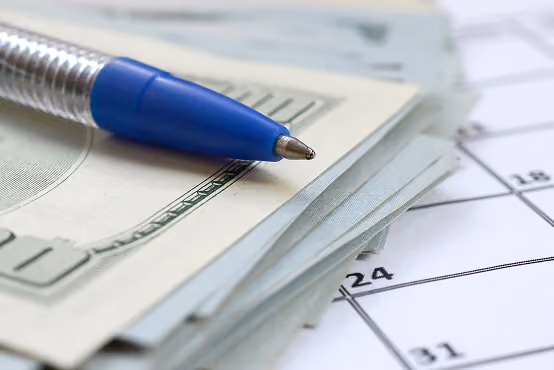 Close-up of a blue pen resting on a stack of US hundred dollar bills atop a calendar page.