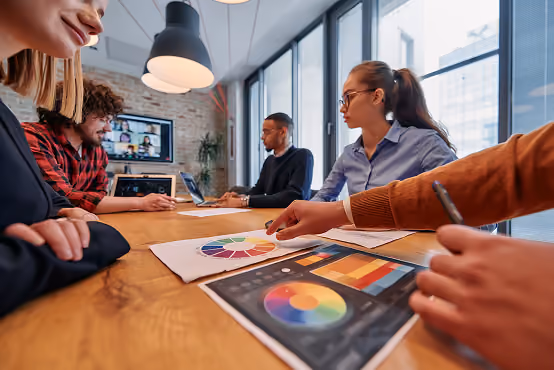 Group discussing color charts and graphics at a conference table in a modern office with video call on screen.